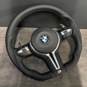 Könnte beinhalten: Schwarzes Lederlenkrad mit perforierten Griffbereichen, roten Nähten und silbernen Akzenten. Mit BMW-Logo und integrierten Bedienelementen. Ein Hochleistungs-Autozubehör.
