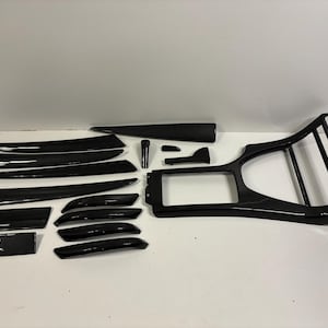 May include: A selection of glossy black car interior trim components, including dashboard elements, console trim, and door panels. The parts feature a carbon fibre weave pattern, suggesting a high-performance aesthetic. The set is arranged on a white surface.