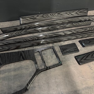 May include: A set of automotive interior trim pieces featuring a glossy black and grey wood grain pattern. The collection includes several long, narrow strips, a large centre console panel with cutouts, and two smaller rectangular accent pieces.