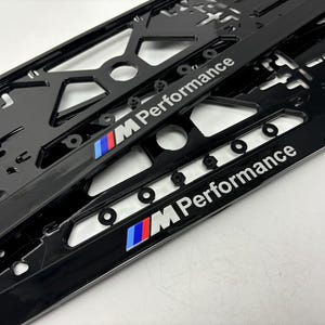 Op de afbeelding: Zwarte kentekenplaathouders met de tekst "M Performance" in het wit. De frames hebben een geometrisch ontwerp en het BMW-logo in blauw, rood en wit. De frames zijn gemaakt van glanzend materiaal.