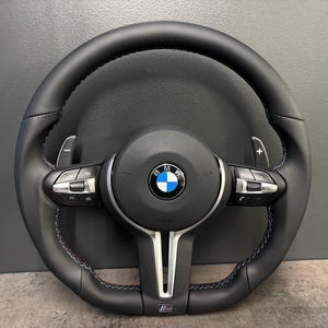 May include: Black leather steering wheel with blue stitching and silver detailing. Includes the BMW emblem and paddle shifters. A sporty car accessory, designed for performance and style.
