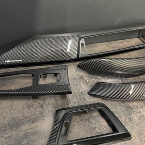 May include: A selection of carbon fibre car interior trim components. The pieces have a glossy finish and a woven pattern. The largest piece has "M Performance" text. The set includes dashboard, console, and door trim.