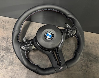 May include: Black leather steering wheel with white stitching and a BMW logo in the center. The wheel has integrated controls and paddle shifters. The M logo is visible at the bottom.