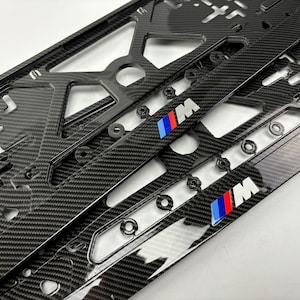 Puede incluir: Un marco para matrícula de fibra de carbono negra con acabado brillante y diseño recortado. El marco muestra el logotipo M con rayas azules, moradas y rojas en la parte inferior derecha y en la barra central, siendo un accesorio automotriz.