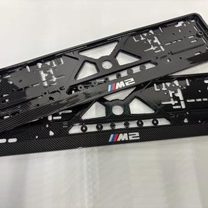 Może przedstawiać: Dwie czarne ramki tablic rejestracyjnych z wzorem z włókna węglowego. Każda ramka ma litery "M2" z czerwonymi, białymi i niebieskimi paskami. Ramki mają geometryczny kształt z różnymi wycięciami i otworami montażowymi.