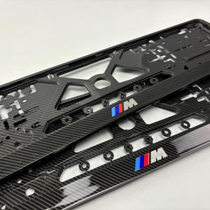 Può includere: Due cornici nere per targa in fibra di carbonio con il logo BMW M in blu, rosso e bianco. Le cornici hanno una finitura lucida e un motivo in fibra di carbonio intrecciata. Progettato per uso automobilistico.