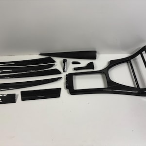 May include: A selection of glossy black carbon fibre car interior trim pieces. The set includes various shapes and sizes, such as long, curved panels, a console frame, and smaller rectangular pieces. The parts are arranged on a white surface.