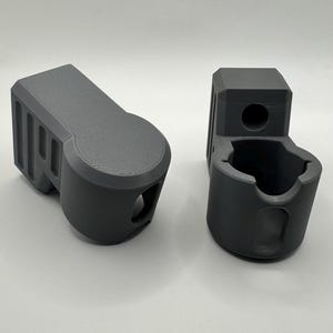 Pode incluir: Dois objetos cinzentos impressos em 3D. Um é retangular com uma extremidade arredondada e ranhuras de ventilação. O outro tem uma base cilíndrica e uma abertura circular. Ambos são feitos de um material cinzento mate e estão sobre uma superfície branca.