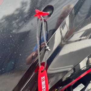May include: A red safety tag with the text "REMOVE BEFORE USE" hangs from a black engine component. The tag is attached with a black cord and a red plastic clip. The engine component has red accents.