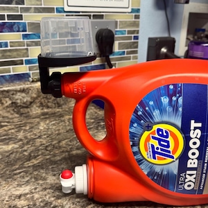 Könnte beinhalten: Eine orangefarbene Tide-Waschmittelflasche mit einem schwarzen Spender und einem durchsichtigen Messbecher. Die Flasche hat ein blau-weißes Etikett mit der Aufschrift "Ultra Oxi Boost". Im Hintergrund befindet sich ein Schild mit der Aufschrift "Wash-Dry-Fold-Press".