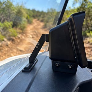 May include: A black electronic display unit mounted on a vehicle dashboard with a sturdy angled support arm. The device features a matte finish, visible silver screws, and a protective housing, positioned to face the driver while overlooking a dirt trail.