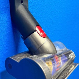 May include: A black and gray Dyson vacuum cleaner attachment. The attachment has a red button and a clear plastic section with the Dyson logo. The attachment is mounted on a blue wall.