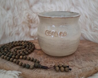 Könnte beinhalten: Beige Keramikbecher mit dem Text "GAIA MA" auf der Seite. Eine Holzperlenkette liegt neben dem Becher auf einer Holzoberfläche. Der Hintergrund ist ein weicher, strukturierter Stoff.
