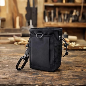 Könnte beinhalten: Eine schwarze Utility-Tasche mit Karabinerhaken und Kordelzugverschluss. Die Tasche hat eine Netztasche an der Seite und einen D-Ring-Befestigungspunkt. Sie ist zum Tragen am Gürtel oder zur Befestigung an einer Tasche konzipiert.