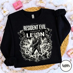 Może przedstawiać: Czarna bluza z grafiką z Resident Evil. Projekt zawiera napis "Resident Evil Requiem Leon" oraz ilustrację postaci trzymającej broń, otoczonej przez zombie i budynki.