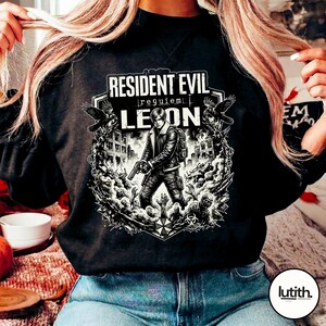 Może przedstawiać: Czarna bluza z grafiką z gry "Resident Evil: Requiem". Projekt zawiera tekst "Resident Evil" i "Leon", a także ilustrację postaci z bronią, otoczonej przez zombie i kruki.