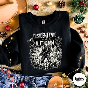 Może przedstawiać: Czarna bluza z okrągłym dekoltem z grafiką Resident Evil. Projekt zawiera tekst "Resident Evil Requiem Leon" oraz ilustrację postaci z bronią, otoczonej przez zombie i budynki w czerni i bieli.