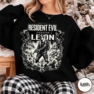 Może przedstawiać: Czarna bluza z grafiką z Resident Evil. Projekt zawiera tekst "Resident Evil | requiem | Leon" oraz ilustrację postaci trzymającej broń, otoczonej przez zombie i budynki.