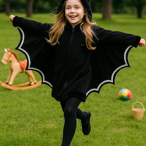 May include: A child wearing a black bat costume with a hood and white trim on the wings, running on green grass. The costume has a zipper down the front. A rocking horse, a colorful ball, and a small bucket are in the background.