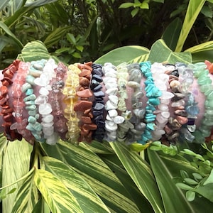 May include: A collection of gemstone chip bracelets in various colors, including red, turquoise, white, and green. The bracelets are displayed on a pink cylindrical form, set against a backdrop of green foliage.