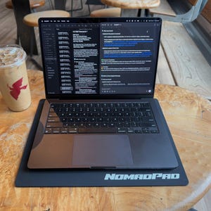 Può includere: Un laptop nero con schermo illuminato che mostra testo è appoggiato su un NomadPad nero. A sinistra c'è un bicchiere di plastica trasparente con caffè freddo e un logo rosso. Il laptop è su un tavolo di legno.
