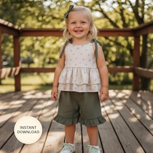 K&ouml;nnte beinhalten: Ein Kind tr&auml;gt ein zweiteiliges Outfit. Das Oberteil ist ein helles Tanktop mit floralem Muster und gr&uuml;nen Tr&auml;gern. Die Shorts sind olivgr&uuml;n mit R&uuml;schensaum. Das Kind tr&auml;gt auch eine passende Schleife und wei&szlig;e Turnschuhe.