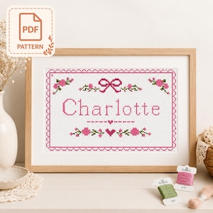 May include: A cross-stitch pattern in a wooden frame featuring the name Charlotte in pink letters, surrounded by a pink decorative border, a pink bow, small pink flowers, and green leaves on a white fabric background with a small heart icon.