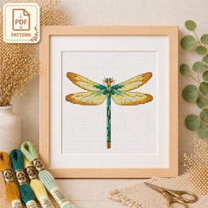 May include: A framed cross-stitch pattern of a dragonfly, featuring yellow, teal, and brown wings and body. The artwork is in a light wood frame. The image includes embroidery floss in various colors and a PDF pattern icon.