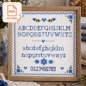 May include: A framed cross-stitch sampler featuring a blue and green alphabet in both uppercase and lowercase letters, along with the numbers 0 through 9. The design includes small blue heart and floral motifs, all enclosed within a decorative border.