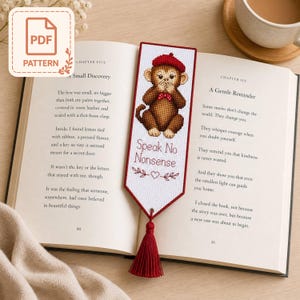 May include: A cross-stitch bookmark with a monkey design, wearing a beret and bow tie, and the text "Speak No Nonsense". It has a red tassel and is placed in an open book. The bookmark is a handmade item.