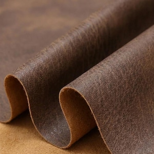 Puede incluir: Primer plano de cuero marrón con una superficie texturizada. El cuero está doblado, revelando una parte inferior marrón más clara. El material tiene un aspecto envejecido, con variaciones de color y tono, lo que sugiere una estética vintage o rústica.