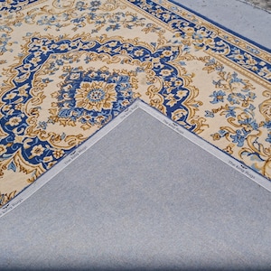 May include: A rectangular area rug featuring a cream-colored background with intricate blue and gold floral patterns and a decorative border. The underside is a plain gray fabric with a white edge tape printed with the text The King Rugs.