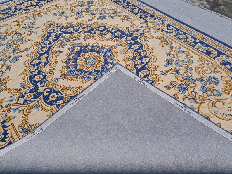 May include: A rectangular area rug featuring a cream-colored background with intricate blue and gold floral patterns and a decorative border. The underside is a plain gray fabric with a white edge tape printed with the text The King Rugs.