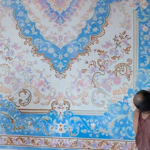 May include: A large rectangular rug featuring a white background with intricate blue and light pink floral patterns. The design includes a central medallion and ornate borders with scrolling vines and blossoms, creating a symmetrical layout.