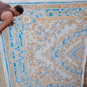 May include: A large rectangular rug featuring an intricate floral and vine pattern in shades of light blue, white, and gold. The design includes symmetrical scrolling motifs and a decorative border, displayed vertically against a brick wall.