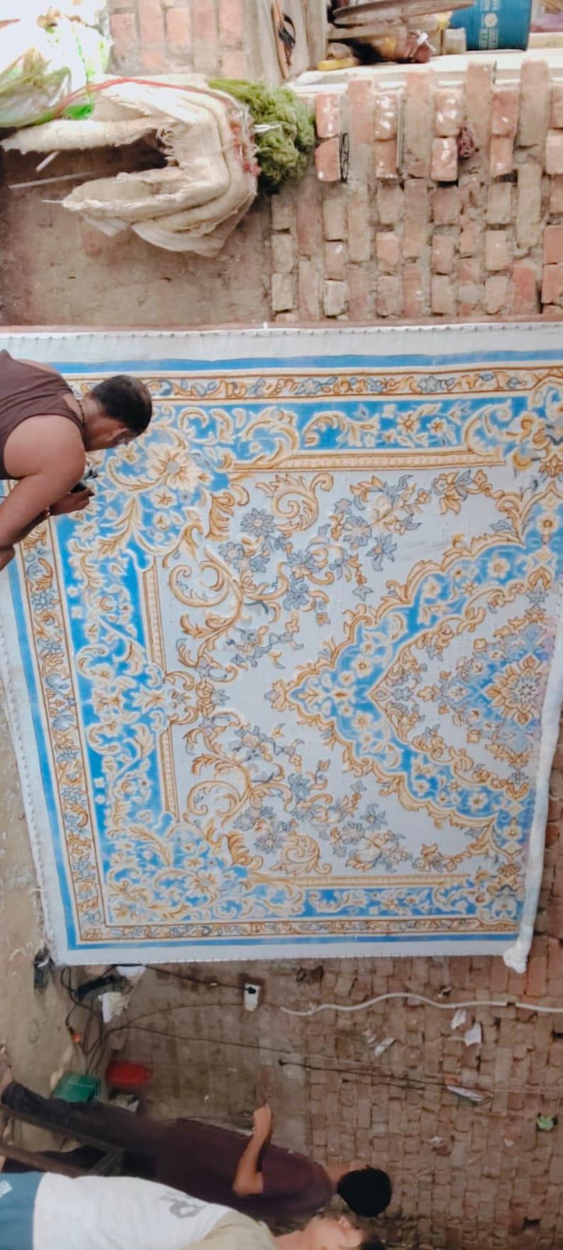 May include: A large rectangular rug featuring an intricate floral and vine pattern in shades of light blue, white, and gold. The design includes symmetrical scrolling motifs and a decorative border, displayed vertically against a brick wall.