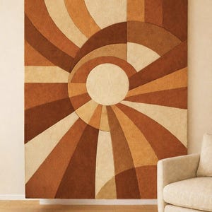 Op de afbeelding: Een groot, rechthoekig vloerkleed met een geometrisch zonnesterontwerp in tinten bruin, beige en crème. Het vloerkleed is aan een muur bevestigd, met een neutraal gekleurde fauteuil in de hoek. Het ontwerp heeft concentrische cirkels en stralende lijnen.