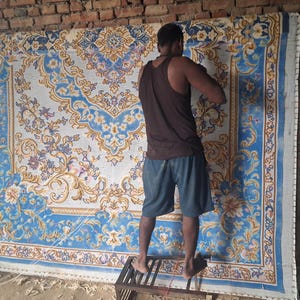 May include: A large rectangular rug featuring an intricate floral and scrollwork pattern in shades of blue, white, and gold. The design includes a central medallion and a decorative border, showcasing detailed craftsmanship on a woven textile surface.