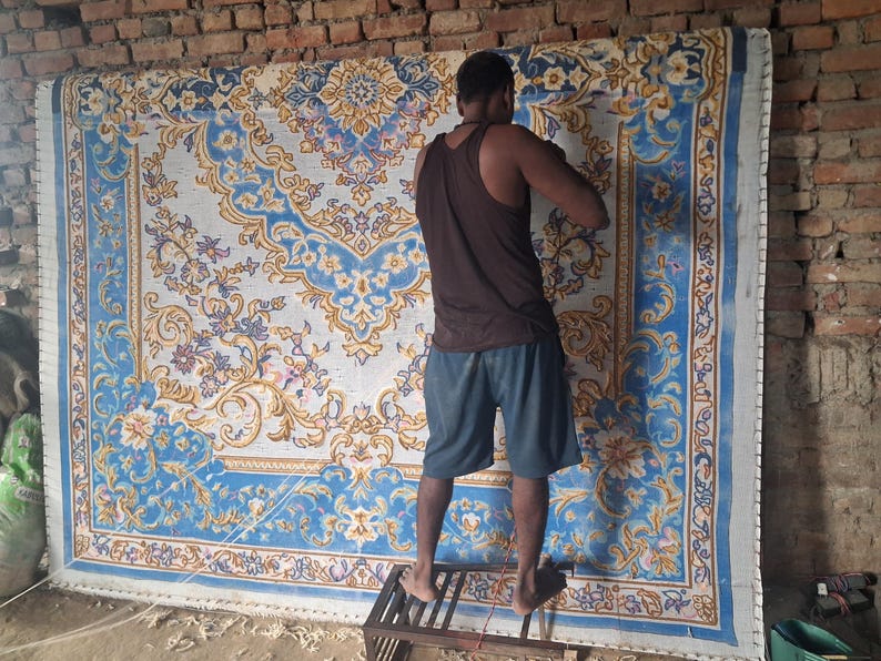 May include: A large rectangular rug featuring an intricate floral and scrollwork pattern in shades of blue, white, and gold. The design includes a central medallion and a decorative border, showcasing detailed craftsmanship on a woven textile surface.