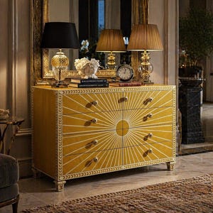 Puede incluir: Una cómoda rectangular de color amarillo mostaza con un patrón de rayos de sol blanco que irradia desde el centro. La pieza tiene ocho cajones con pequeños pomos redondos, un borde decorativo en los laterales y se apoya sobre cuatro patas talladas.