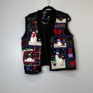 Könnte beinhalten: Eine schwarze ärmellose Weste mit einem Patchwork-Design und gestickten Weihnachtsmotiven wie Schneemännern, roten Fäustlingen, Geschenken und einem Herz. Die Weste hat eine Knopfleiste und ein schwarzes Innenfutter mit einem kleinen Logo.