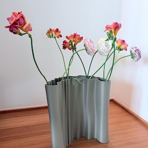Op de afbeelding: Een moderne, grijze vaas met golvende randen bevat een verzameling kleurrijke bloemen. De bloemen omvatten oranje, roze en witte bloemen met groene stelen. De vaas staat op een houten vloer tegen een witte muur.