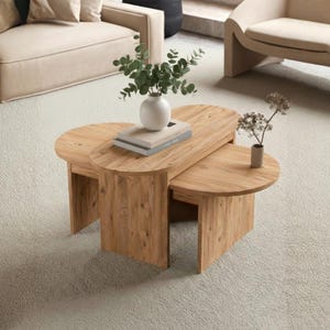 Puede incluir: Una mesa de centro de madera clara con un diseño único e entrelazado. La mesa presenta tres secciones, con un óvalo central y dos mesas laterales semicirculares. Un jarrón blanco con vegetación y libros encima.