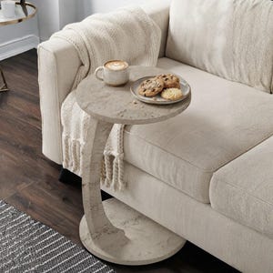 May include: A C-shaped side table with a light beige stone-like finish. The round tabletop holds a speckled ceramic mug filled with a latte and a small plate containing three chocolate chip cookies. The table features a curved pedestal base.