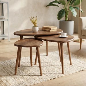 May include: A set of three nesting coffee tables made of dark brown wood with smooth, rounded, organic-shaped tops and tapered legs. The tables are arranged in a tiered formation, with one small white vase and a stack of books placed on top.