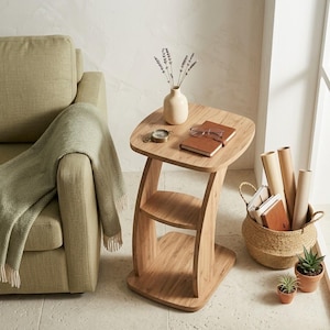 May include: A light oak side table with a curved design, featuring a top shelf and a lower shelf. The table holds a small vase with lavender, a compass, a notebook, and glasses. A woven basket with books and plants sits nearby.