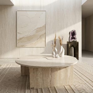 May include: A modern living room featuring a large, round, beige stone coffee table. Two white vases with dried plants sit on the table. A large abstract painting hangs on the wall, and a rug is on the floor. A large amethyst geode sits on a dark brown stand.