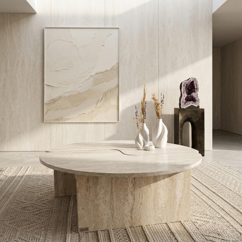 May include: A modern living room featuring a large, round, beige stone coffee table. Two white vases with dried plants sit on the table. A large abstract painting hangs on the wall, and a rug is on the floor. A large amethyst geode sits on a dark brown stand.