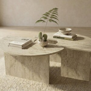 May include: A two-piece, light-colored stone coffee table with a rounded design. The table features a small cactus in a decorative pot, books, and a small bowl. A fern leaf adds a touch of greenery to the minimalist decor.