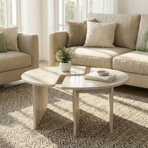 May include: A light-colored, round coffee table with a unique cutout design. The table has a light wood finish and is supported by three legs. A small potted plant and books are on the table, which sits on a patterned rug.